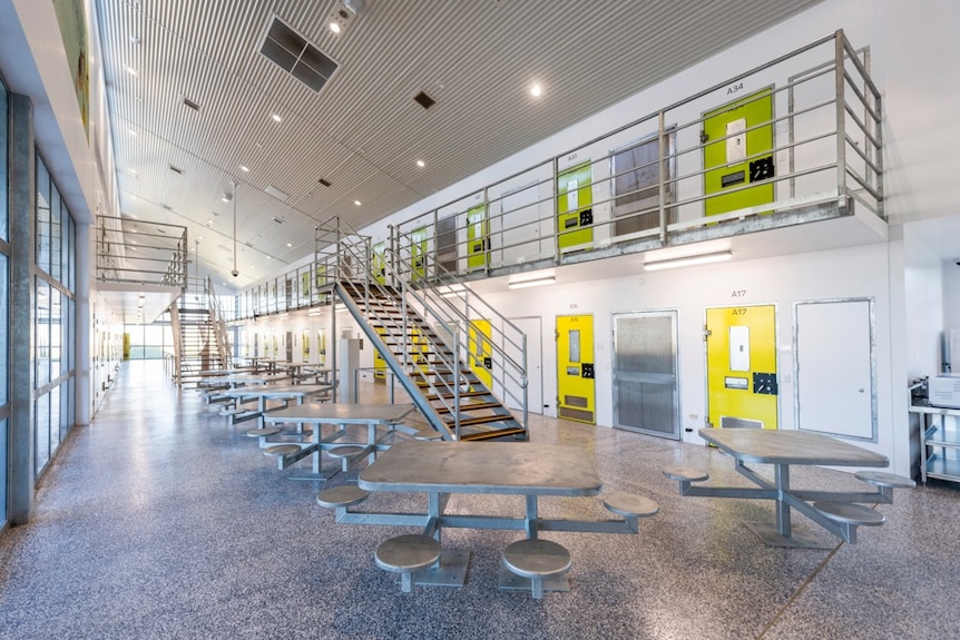 Interior view of a newly-built prison with shiny new yellow doors on cells and metal dining table along a hallway.