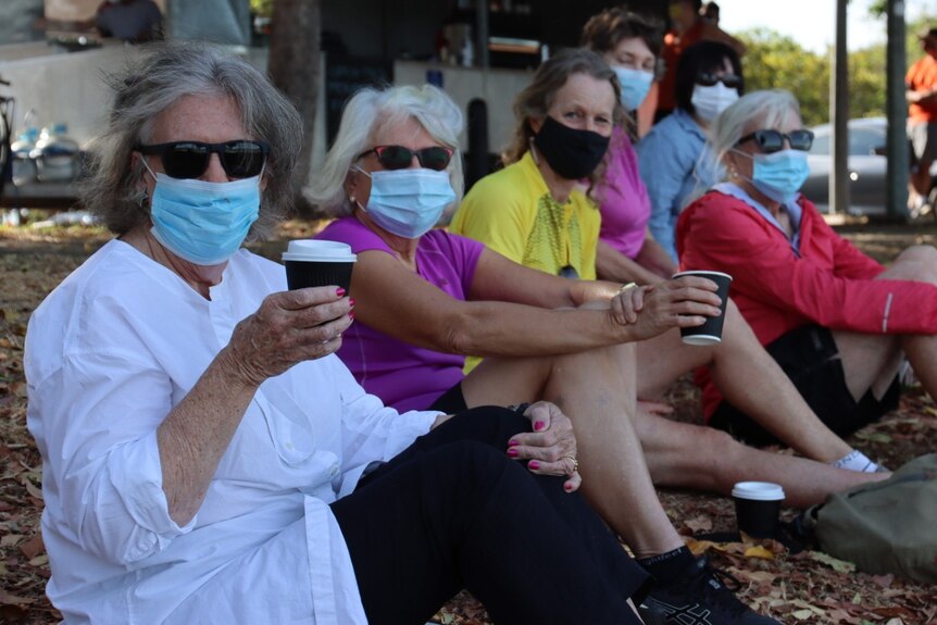 A group of masked women sit in an area in coastal Darwin, wearing masks and holding coffee cups.