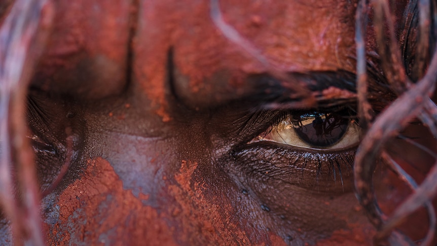 Colour square cropped super close-up image of a man's face, his skin painted-up in red Pindan country dirt.