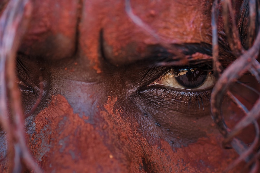 Colour square cropped super close-up image of a man's face, his skin painted-up in red Pindan country dirt.