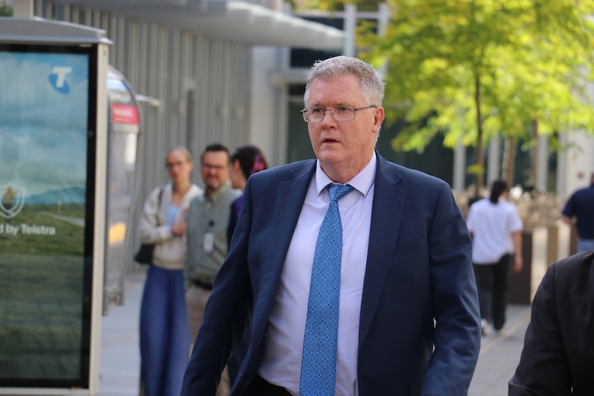 A man wearing a blue blazer, white shirt and blue tie and glasses walks along a busy street.
