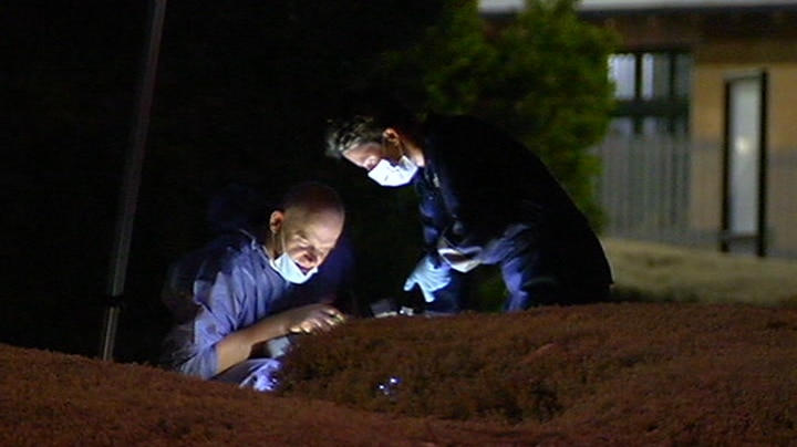 Two people in blue overalls and face masks work in the dark with flashlights under an emergency services tent.