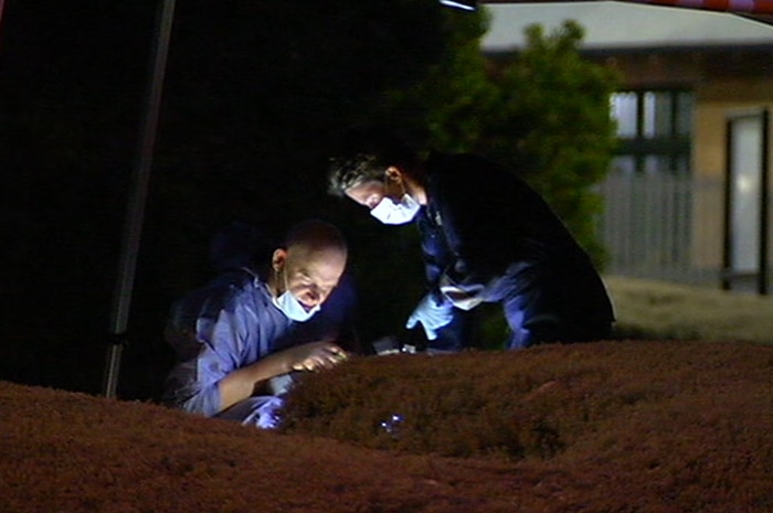 Two people in blue overalls and face masks work in the dark with flashlights under an emergency services tent.