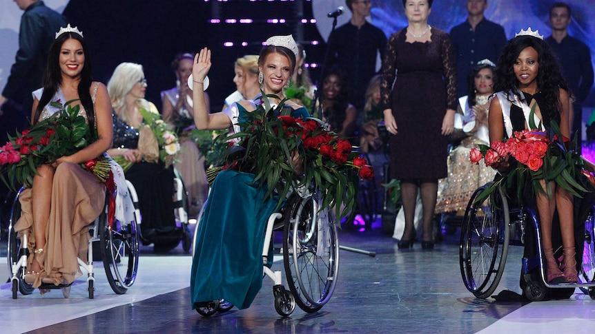 Three beauty pageant contestants in wheelchair smile on stage.