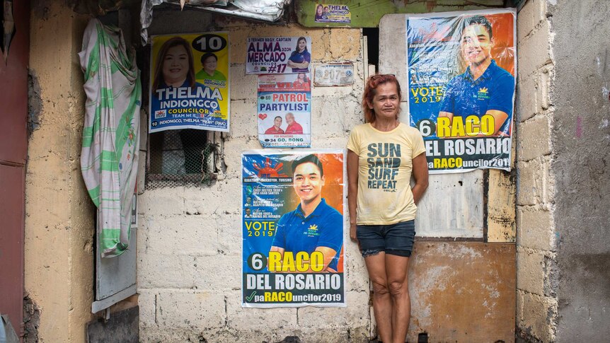 A thin, tired looking woman stands against a concrete wall covered in candidate posters