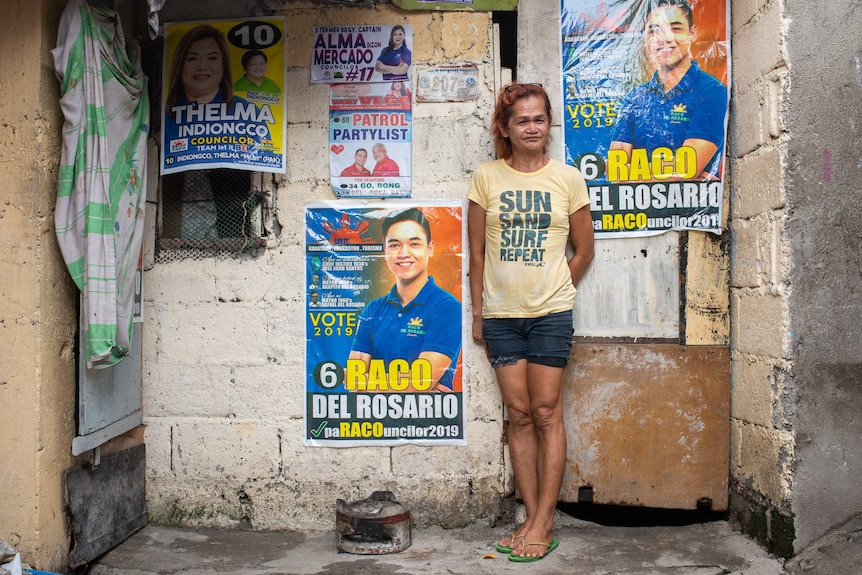 A thin, tired looking woman stands against a concrete wall covered in candidate posters