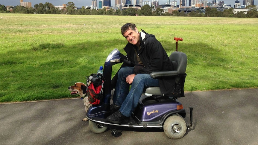 Sean Cox sits in his mobility scooter next to his dog in a green park with the Melbourne skyline in the background.