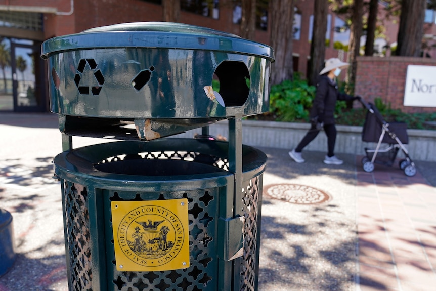 A close-up of a round, dark green metal rubbish bin with ornate designs. A masked woman walks behind it pushing a stroller.