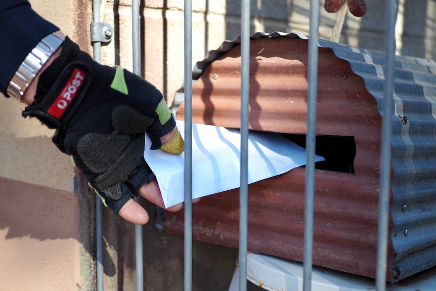 A gloved hand of a postman pokes a letter through a barred fence, slotting it into a corrugated iron mail box