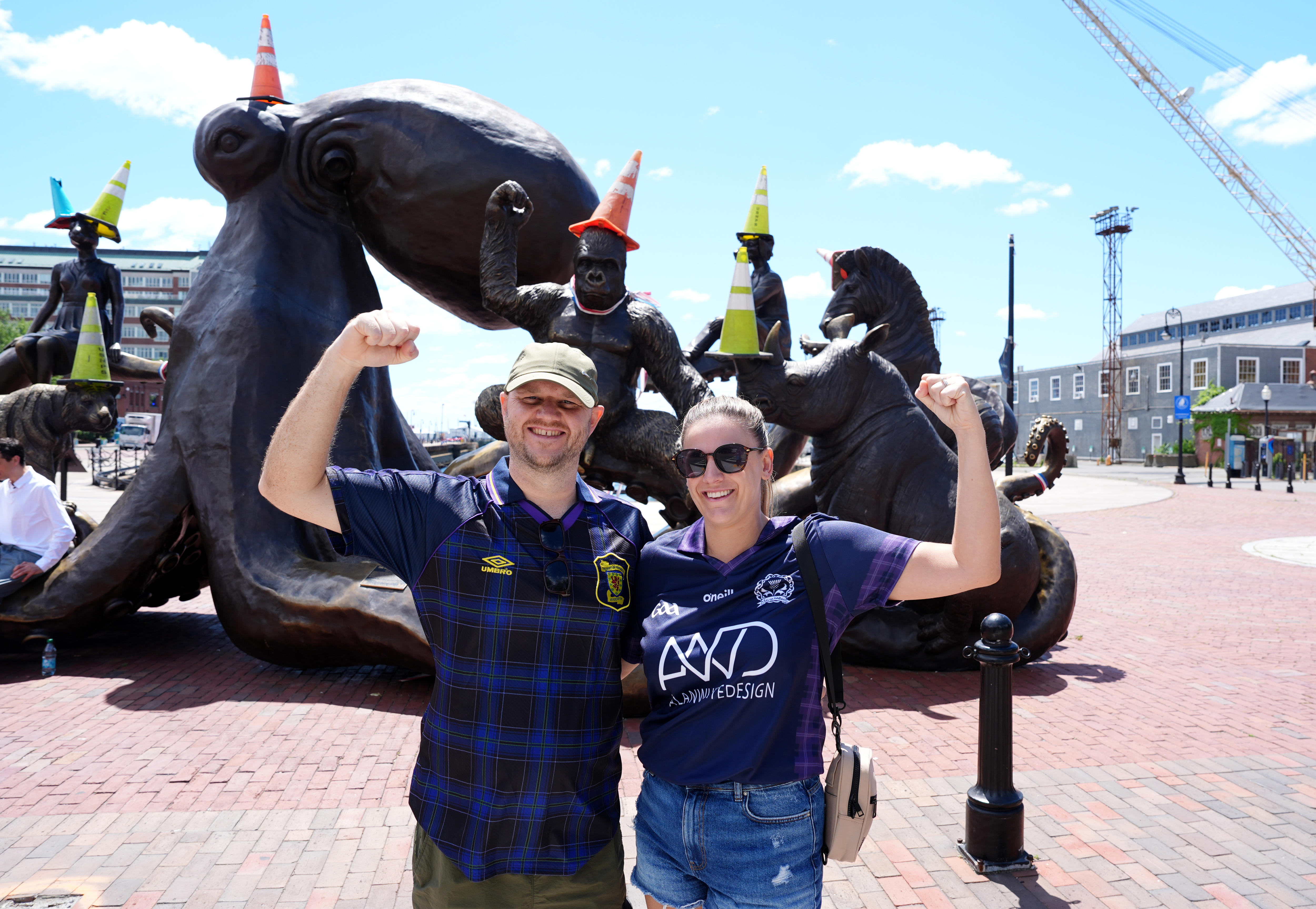Two Scotland fans pose in front of the Arms of Friendship Statue that has traffic cones on it