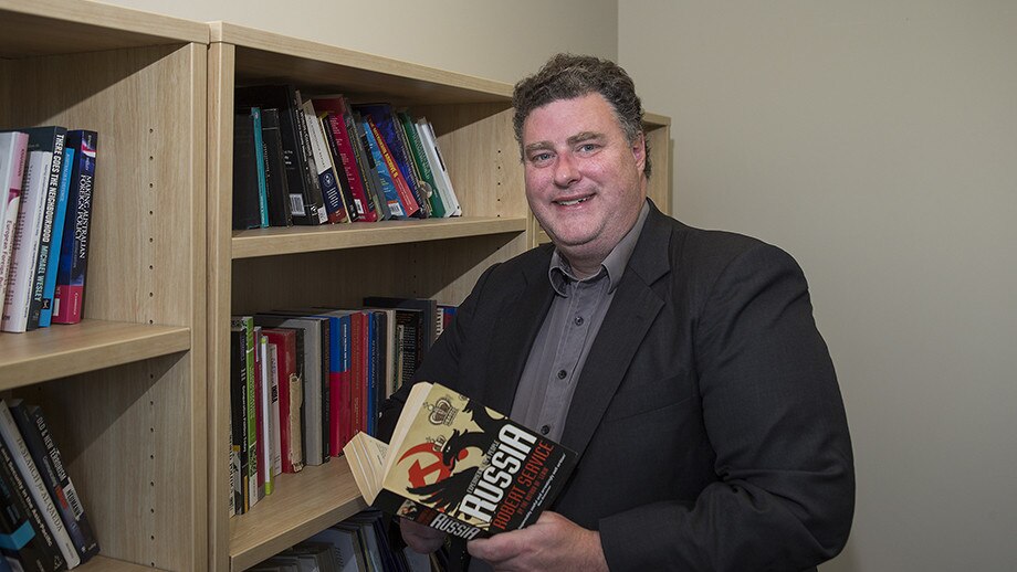 Matthew Sussex, a man with short brown hair dressed in a dark jacket and shirt, stands next to a bookshelf holding a book