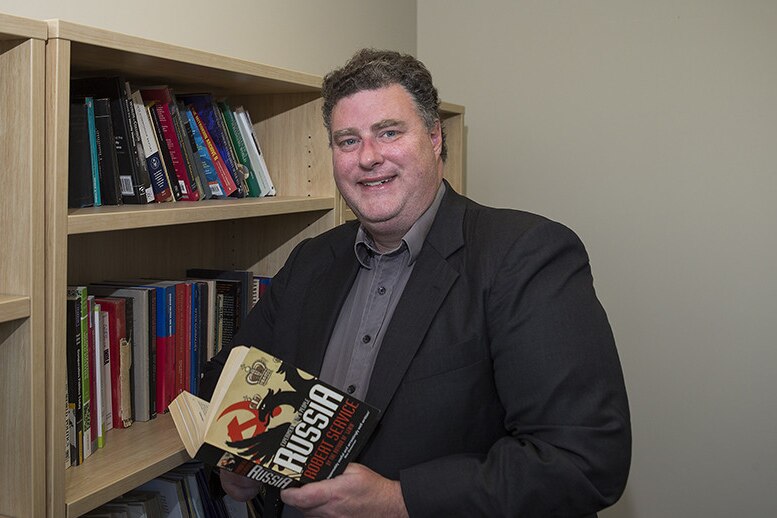 Matthew Sussex, a man with short brown hair dressed in a dark jacket and shirt, stands next to a bookshelf holding a book
