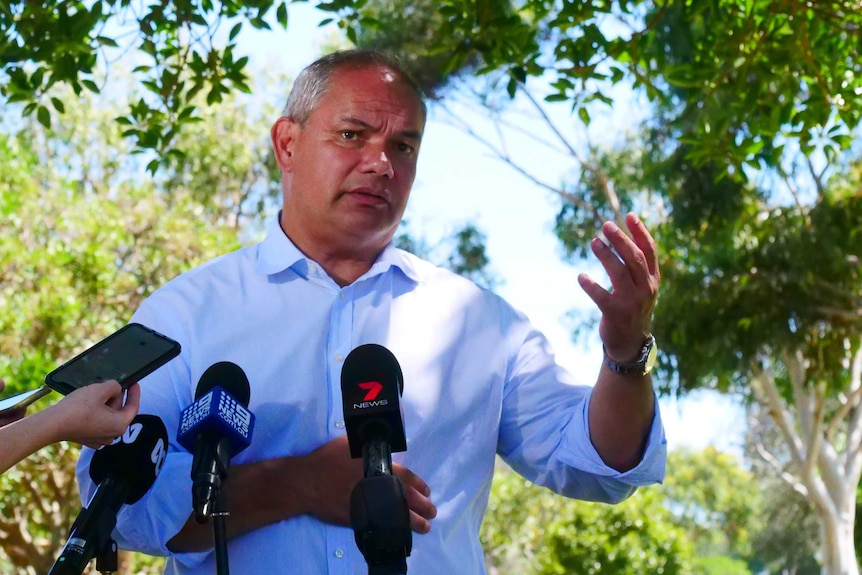 grey-haired, balding man at media conference, wearing a blue shirt with sleeves rolled up and his left arm outstretched