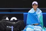 Ash Barty sits in her chair, next to her tennis gear, on-court at the Australian Open, wrapped in towels.