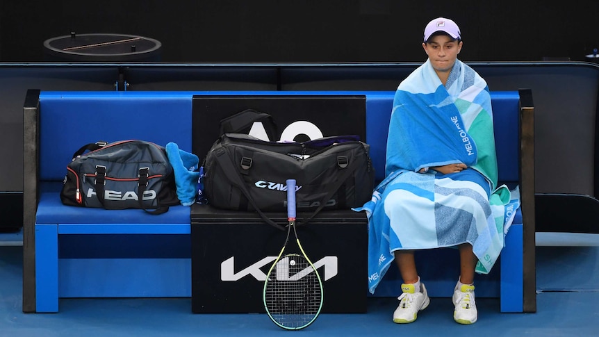 Ash Barty sits in her chair, next to her tennis gear, on-court at the Australian Open, wrapped in towels.