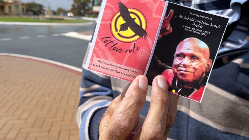 A hand holding a memorial card with a picture of Uncle Archie Roach smiling and pointing.