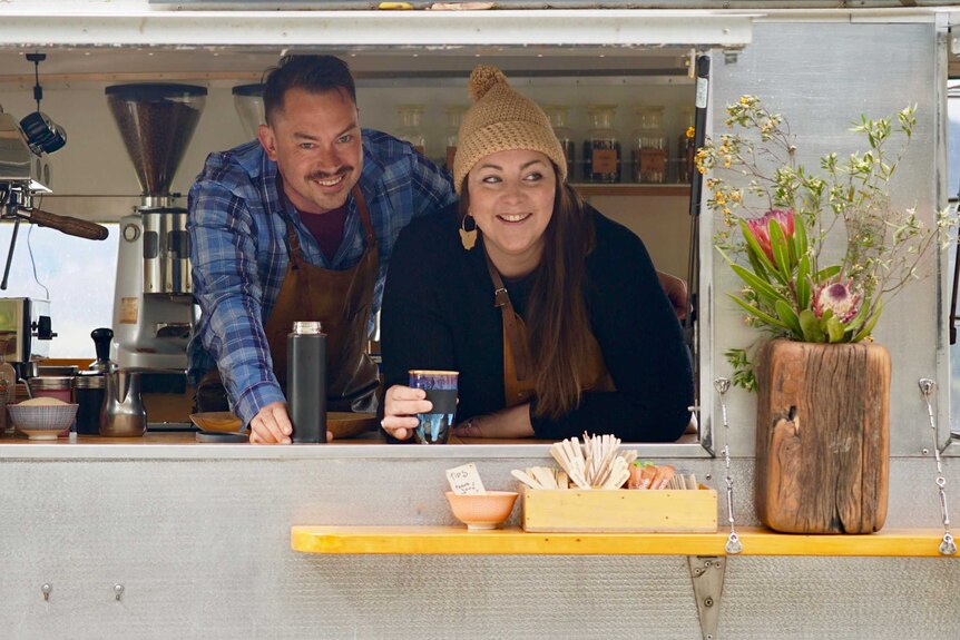 A man and woman smile as they hold coffee cups and peer out of a coffee van.