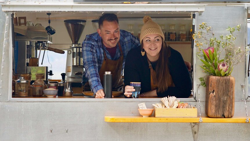 A man and woman smile as they hold coffee cups and peer out of a coffee van.