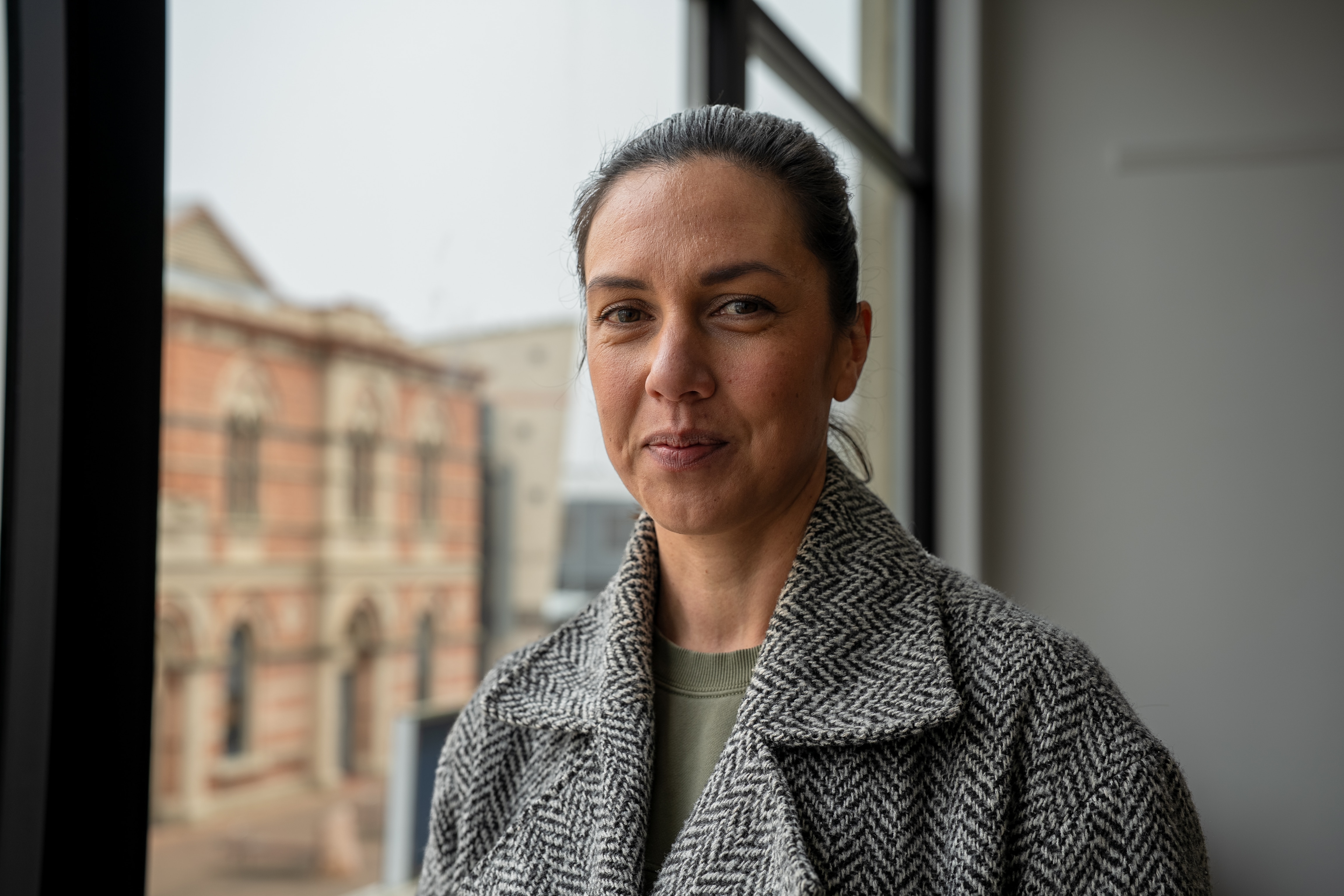 a woman in a grey coat and green shirt standing near a window