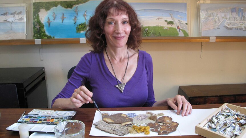 The author/artist sitting at a table holding a paintbrush over an abstract collage. Images from her books are behind her.