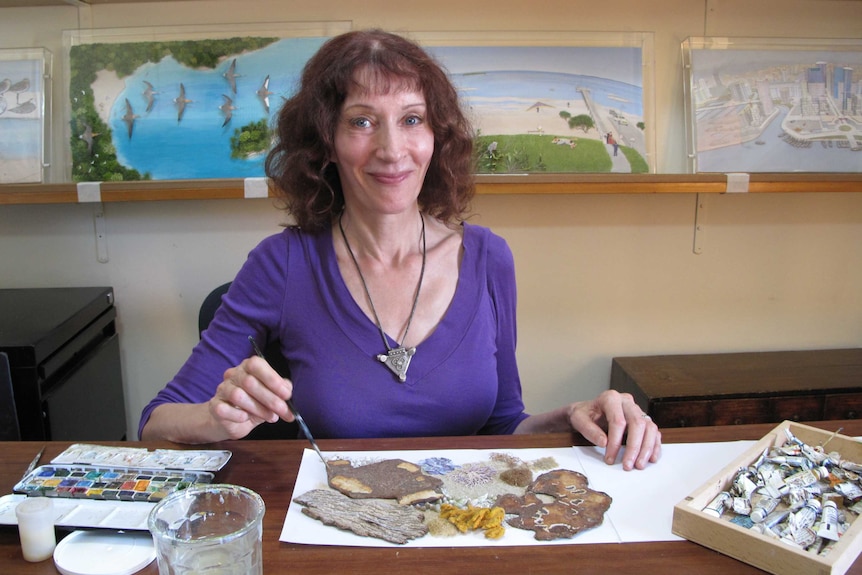 The author/artist sitting at a table holding a paintbrush over an abstract collage. Images from her books are behind her.