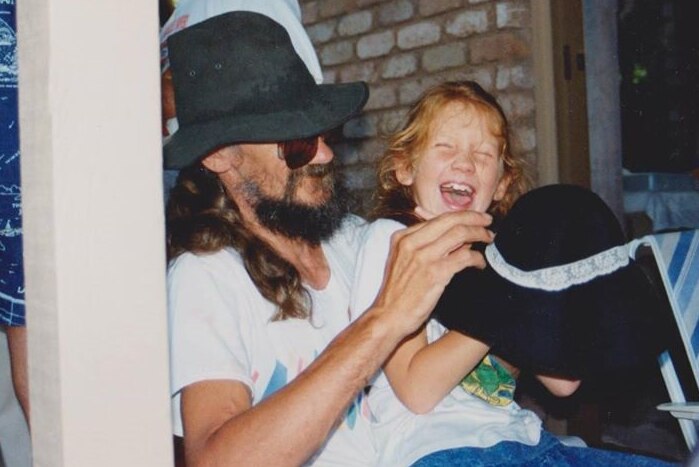 A man with long hair, a beard and wearing a black hat smiles as a young girl with red hair laughs beside him.