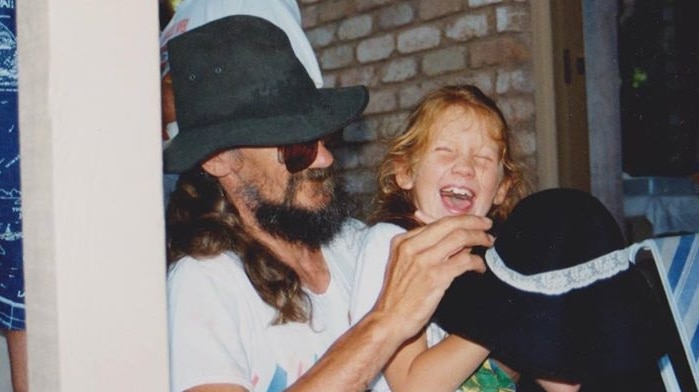 A man with long hair, a beard and wearing a black hat smiles as a young girl with red hair laughs beside him.
