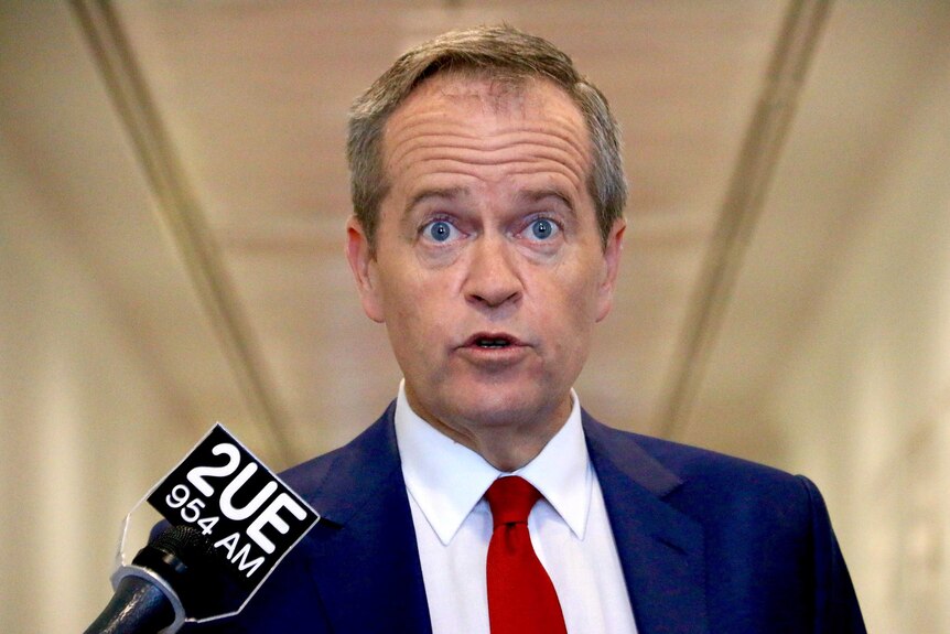 Low front on upper body view of Bill Shorten raising his eyebrows as he speaks with reporters, not in view.