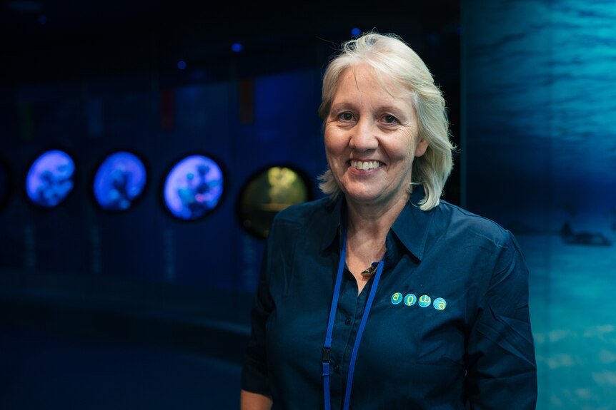 Catherine wears a blue work uniform shirt, standing in front of an aquarium, smiling at the camera.