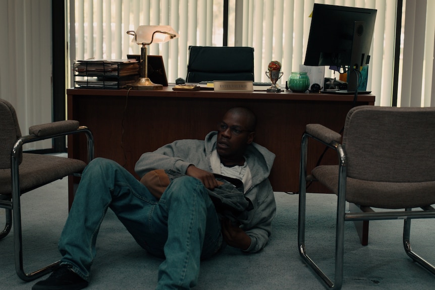 Movie still: Man holding backpack hides under office desk.