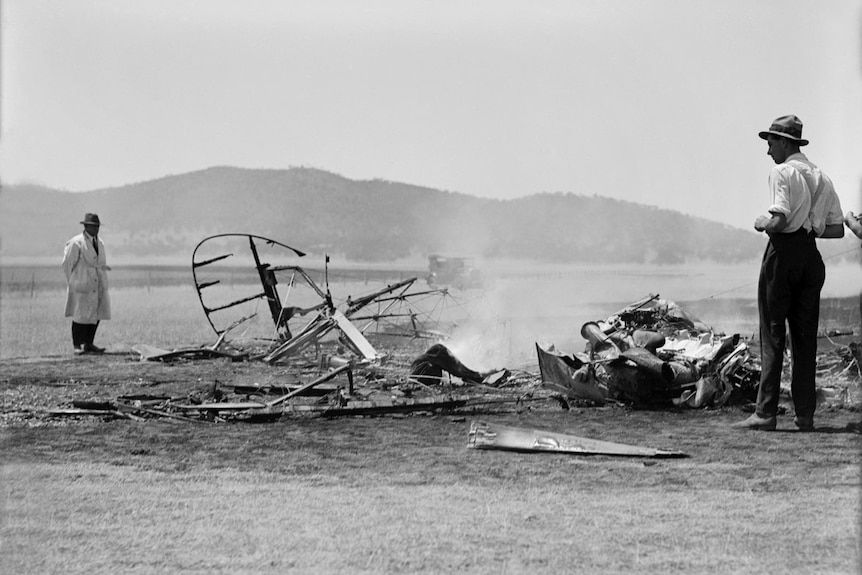 Black and white photo of a plane crash. The plane has been reduced to rubble, which is still smoking, in a field.