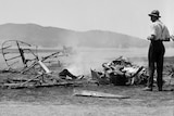 Black and white photo of a plane crash. The plane has been reduced to rubble, which is still smoking, in a field.