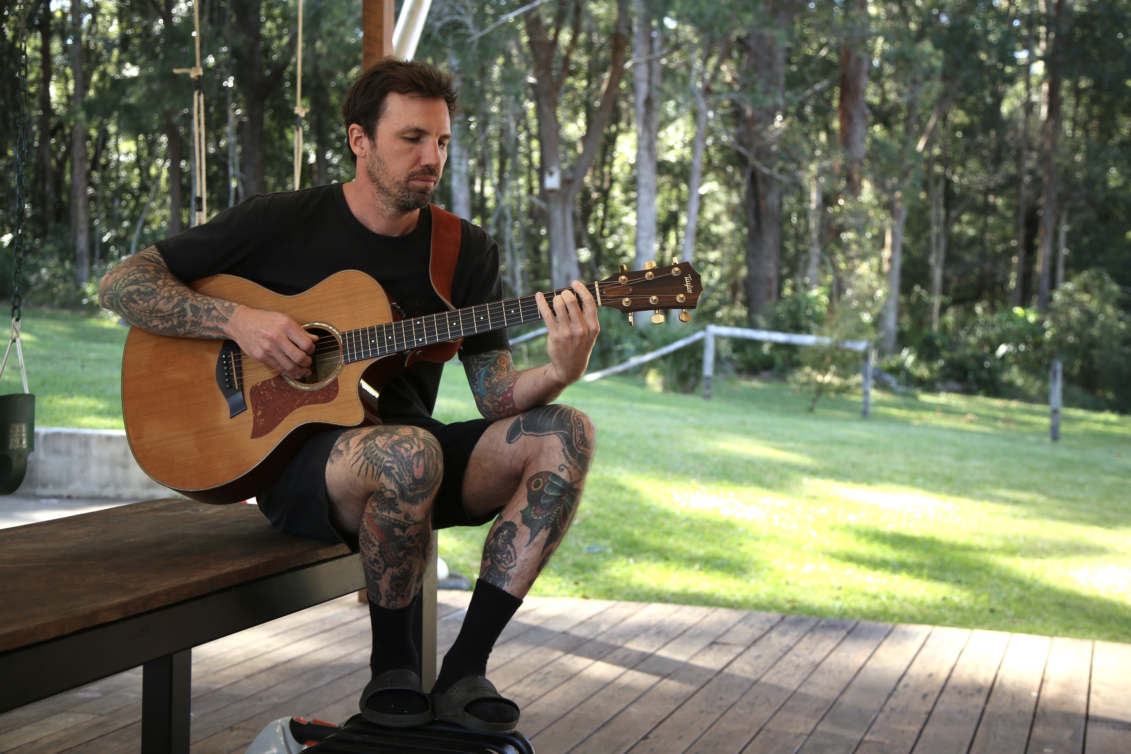 A man in t-shirt and shorts and sandals plays an acoustic guitar on a deck
