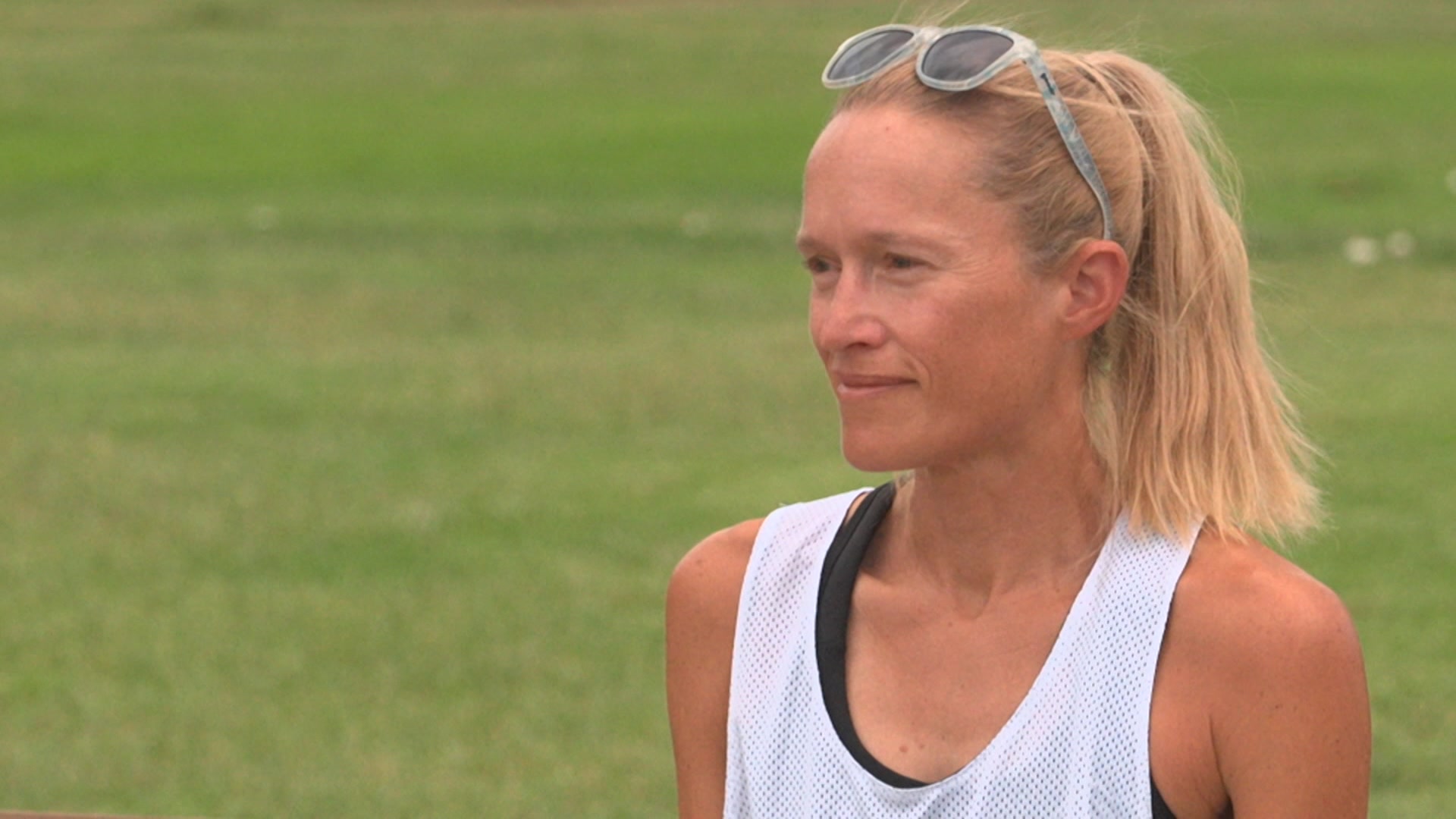 Uma mulher com rabo de cavalo loiro usa uma camiseta branca e óculos escuros na cabeça.