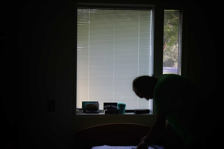 A male patient picks up things from a bed in a bedroom. He is in silhouette and his face has been blurred.