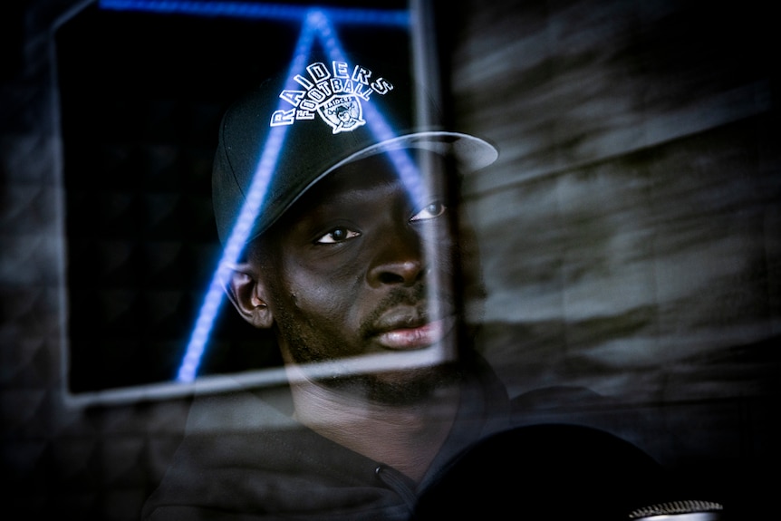 A close up of a man's face, through the glass window of a recording booth. The room he's in is dark.