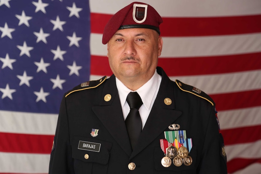 Man wearing military uniform stand in front of US flag.