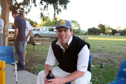 A man in a blue and yellow cap and cricket whites sits with a beer bottle near a cricket oval.