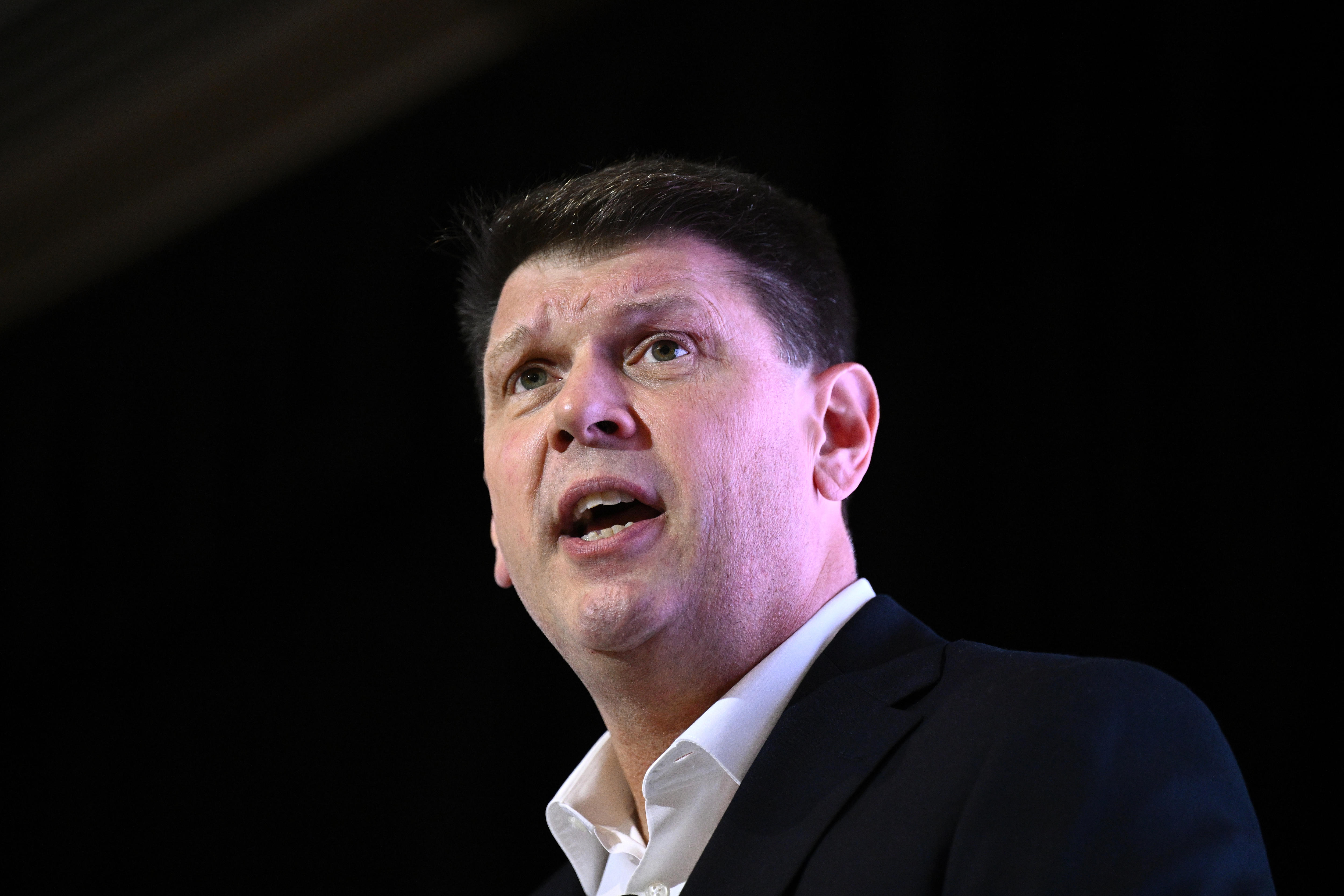 A dark-haired man in a dark suit in mid-sentence.