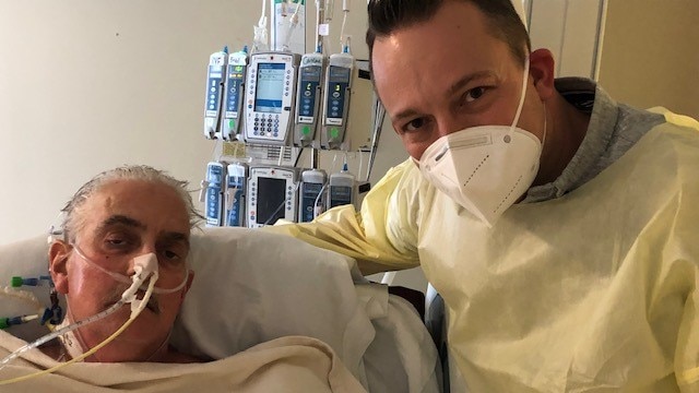 An older man lying in a hospital bed. A younger man wearing a face mask stands beside him