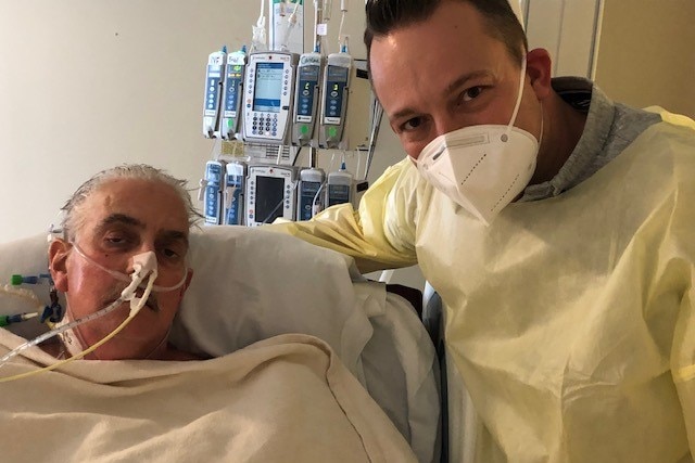 An older man lying in a hospital bed. A younger man wearing a face mask stands beside him