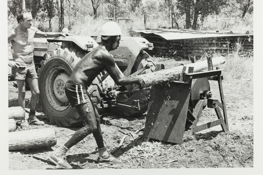 A man wearing only boots, shorts, and a hard hat operating an open air sawmill.