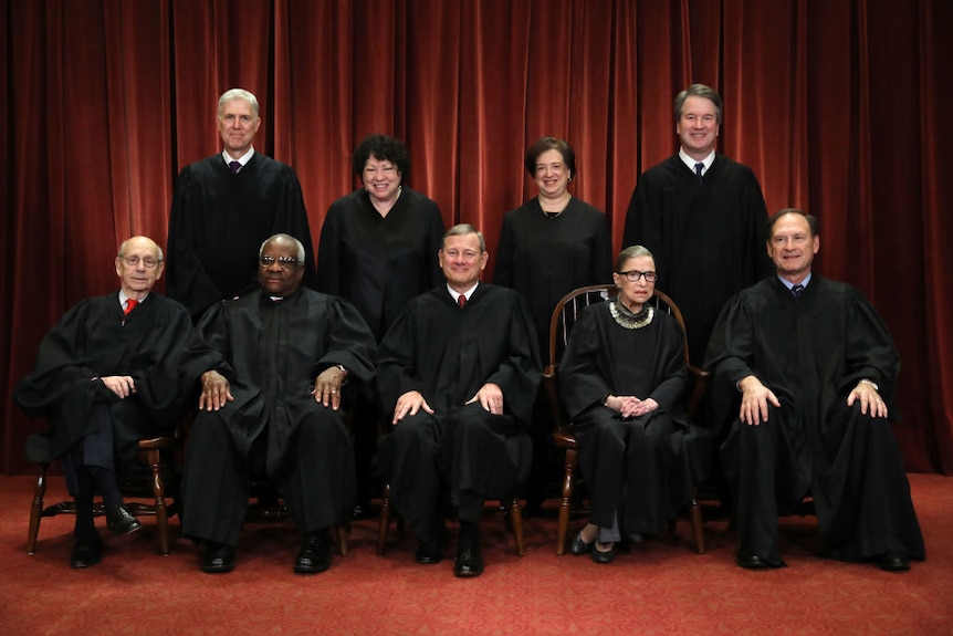 Against a red backdrop in an ornate room 9 judges in court dress pose for a photo
