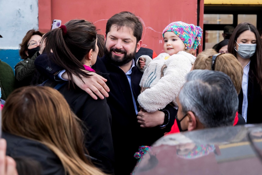 Chilean President Gabriel Boric is holding a baby in his left arm, while hugging a female supporter with his right arm, smiling.