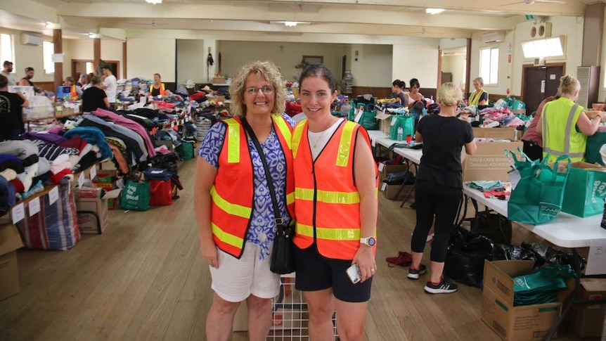 Two women in high-vis vests stand in a busy hall filled with boxes and bags of clothes.