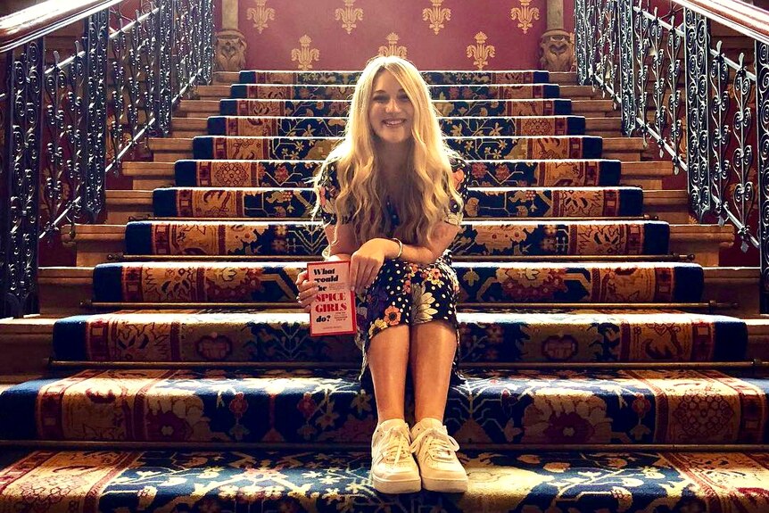 A young blonde woman sits on a grand staircase, smiling and clutching a pink book titled What Would The Spice Girls Do