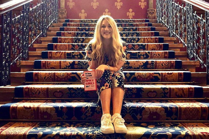 A young blonde woman sits on a grand staircase, smiling and clutching a pink book titled What Would The Spice Girls Do
