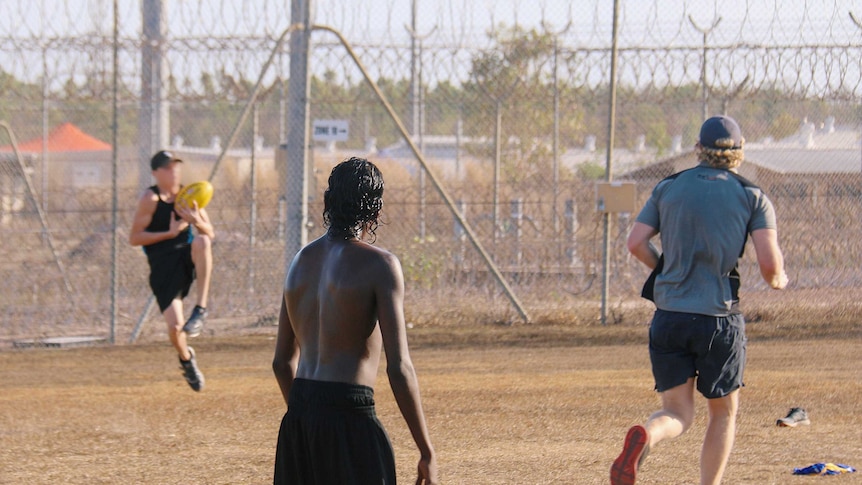 A young indigenous man, with his back to camera, watches on as another young man in a cap takes a mark in a game of football.
