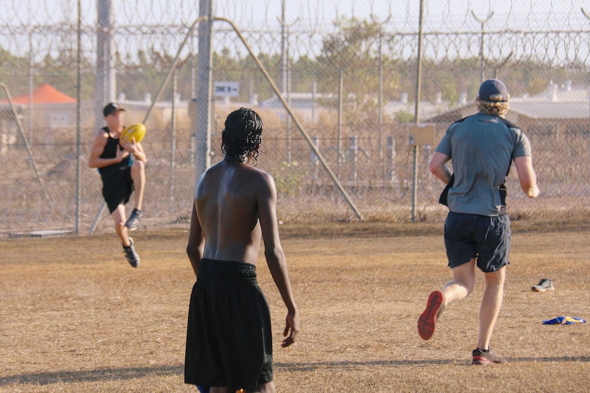A young indigenous man, with his back to camera, watches on as another young man in a cap takes a mark in a game of football.