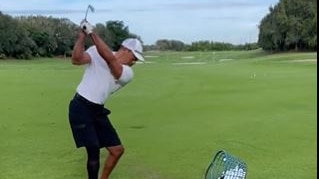 A golfer stands on a driving range with golf balls at his feet, taking a swing with a wedge.